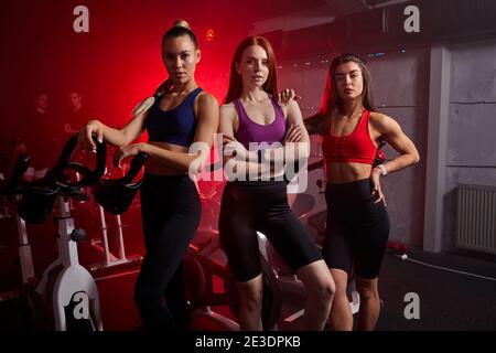 Drei fit Sportlerinnen stehen zusammen in der Nähe von stationären Bikes im Fitnessstudio, posieren vor der Kamera. Isoliert in rot neon beleuchteten Raum im Hintergrund Stockfoto