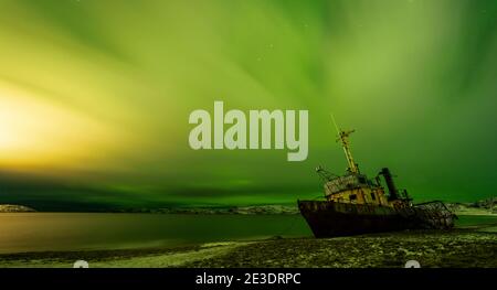 Verlassene Boot auf dem Barentssee Strand in Teriberka, Russland. Ein wolkigen Himmel, der von den Nordlichtern beleuchtet wird. Stockfoto