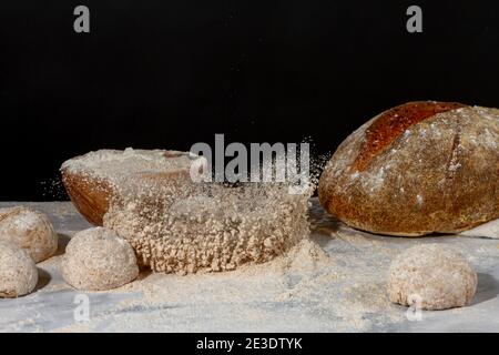 Ein Brotlaib spritzt auf einen Haufen Vollkornmehl auf Marmor-Arbeitsplatte mit dunklem Hintergrund. Es gibt frisches Sauerteig Brot, eine hölzerne Schüssel mit Mehl und Stockfoto