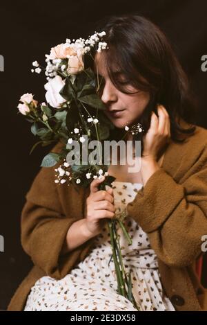 Schöne junge Frau mit Rosen und Blumen auf dunkelschwarzem Hintergrund. Geschlossene Augen und Hand berühren Gesicht. Geschlossene Augen, authentische entspannte Ausdruck A Stockfoto