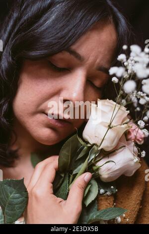 Nahaufnahme der schönen jungen Frau mit Frühlingsblumen und Rosen auf dunkelschwarzem Hintergrund. Geschlossene Augen und Hand berühren Gesicht. Geschlossene Augen, authentische r Stockfoto