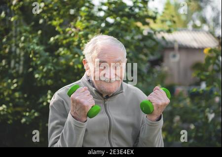 Alter Mann mit Hanteln. Die Fitness des im Garten beschäftigten Menschen. Aktives Alter, gesunder Lebensstil. Stockfoto