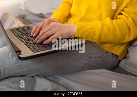 Weibliche Hände tippt Text auf einem Laptop, der auf dem Bett sitzt. Arbeiten Sie von zu Hause Trendfarben von 2021 - Beleuchtung gelb ultimative graue Farbe Stockfoto