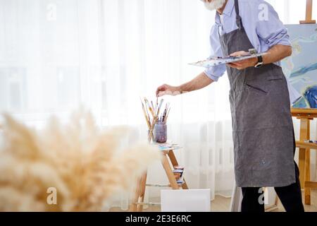 Maler Mann in Schürze unter Pinsel aus der Dose, in hellen Raum. Cropped artsit hält Farben Palette in den Händen Stockfoto