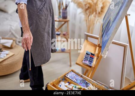 Männlicher Maler Mann in Schürze während des Arbeitsprozesses, männlich mit Pinsel, verschiedene Materialien Farben Werkzeuge für die Malerei Stockfoto