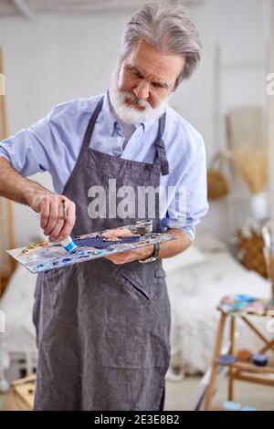 Senior grauhaarige Künstler Mann Malerei mit Maler-Palette und Leinwand, zu Hause, Arbeit als Hobby Stockfoto
