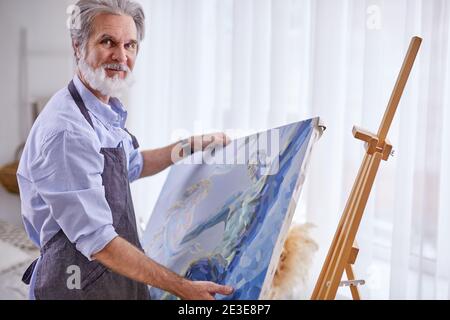 Senior Künstler entfernen Sie die Leinwand von der Staffelei, vorbereitete Malerei in Händen von erfahrenen Künstler. Im hellen Raum Stockfoto