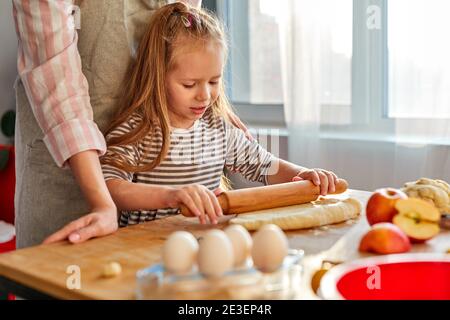 Die junge Mutter lehrt ihre fleißige Tochter zu kochen, steht hinter dem Tisch und rollt den Teig aus Stockfoto