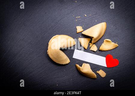 Glückskekse auf einem schwarzen Stein Hintergrund mit einem kleinen roten Herzen. Ein gebrochener Cookie offenbart eine schöne Botschaft. Sehnen Sie sich danach, Ihre zu lesen? Stockfoto