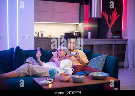 Mann spielt auf Spielkonsole mit Joystick, während Frau Smartphone mit Gaming-App. Millennial Paar lag auf einer Couch zu Hause und spielen Videospiele Stockfoto
