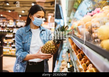 Frau in Schutzmaske und Denim Jacke kauft und Ananas Im Geschäft Stockfoto
