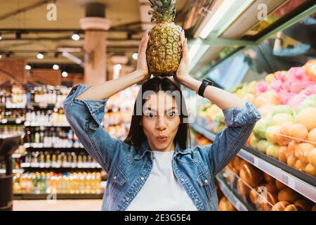 Fröhliche junge Frau in Denim Jacke posiert mit Ananas über Geht in den Laden Stockfoto