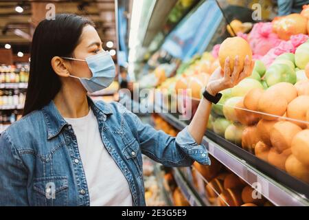 Junge Frau in Schutzmaske und Jeansjacke kauft Obst Im Geschäft Stockfoto