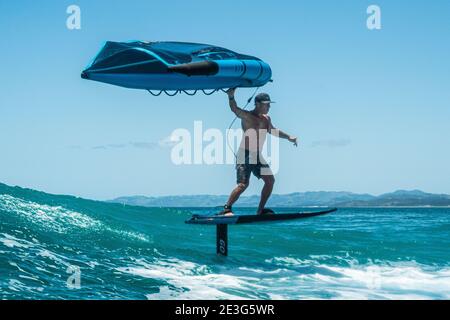 Ein Mann in Shorts reitet eine tropische türkisfarbene Welle mit Tragflügelfolie an einem Sommertag, klaren blauen Himmel. Stockfoto