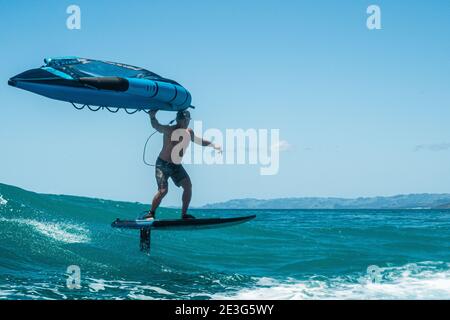 Ein Mann in Shorts reitet eine tropische türkisfarbene Welle mit Tragflügelfolie an einem Sommertag, klaren blauen Himmel. Stockfoto
