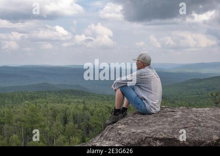 Ein freier Mann mittleren Alters, sitzt auf der Spitze des Berges und genießt eine schöne Aussicht auf das Bergtal. Reisekonzept. Einsamkeit, Einheit mit der Natur. Stockfoto