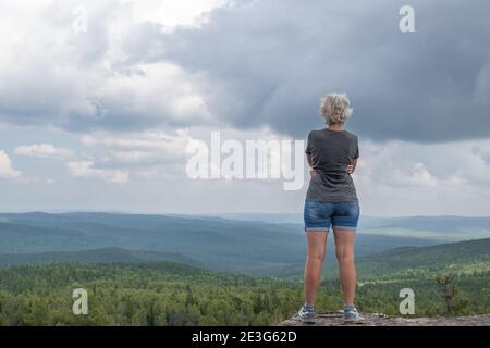 Eine Frau mittleren Alters auf dem Gipfel des Berges genießt einen atemberaubenden Blick auf das hügelige Tal. Freiheit, Ruhe und Entspannung. Gesunder Lebensstil. Stockfoto