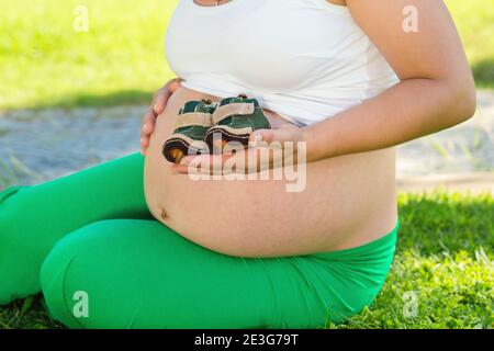 Unerkennbare schwangere Frau berührt ihren Bauch, Bauch spielen mit kleinen Baby-Schuhe sitzen im Freien auf einer grünen Wiese in einem Park auf sonnigen Stockfoto