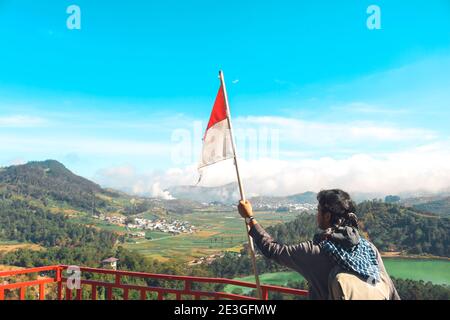 Ein Mann mit indonesischer Flagge, der auf einer Klippe steht und den Blick auf die Natur von See und Berg in Dieng Indonesia genießt. Stockfoto