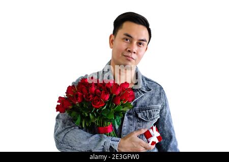 Asiatischer Mann trägt Jeans Jacke mit einem Strauß von rot rosen und rote Geschenkbox isoliert in weißem Hintergrund für Jubiläums- oder Valentinstag-Konzept Stockfoto