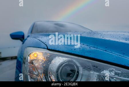 Vorderansicht des blauen Luxus-SUV-Auto in der Nähe von Strand am regnerischen Tag mit Regenbogen als Hintergrund geparkt. Regentropfen auf blauer Textur der Motorhaube. Auto geöffnet Stockfoto