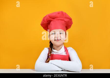 Porträt eines niedlichen kleinen Mädchen Koch auf einem gelben Hintergrund. Das Kind sitzt am Tisch und schaut auf die Kamera. Stockfoto