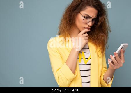 Porträt von nachdenklichen ernst attraktive junge Frau mit lockigen dunkel Blonde Haare tragen gelbe Jacke und optische Brille isoliert auf Hintergrund Stockfoto