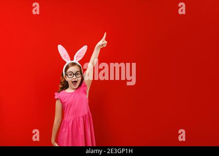 Happy niedlichen kleinen Mädchen tragen osterhasen Ohren und Brille auf rotem Hintergrund. Das untergeordnete Element zeigt auf einen leeren Kopierbereich. Stockfoto