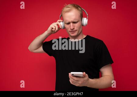 Verärgert nachdenklich gut aussehende blonde junge Mann trägt schwarzes T-Shirt und Weiße Kopfhörer stehen isoliert auf rotem Hintergrund mit Kopierplatz Halten Stockfoto