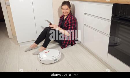 Positive Frau mit Smartphone sitzt auf dem Boden Blick auf zeitgenössische Roboter-Staubsauger, der Schmutz vom Holzboden in der Küche entfernt Zu Hause Stockfoto