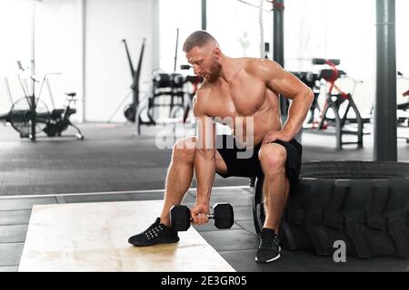 Gym Athlet Bicep Übung Hantel muskulösen Mann sitzen Rad halten Lift Hantel. Funktionelles Cross-Training in Innenräumen. Gut aussehend kaukasisch bärtig Kerl tun w Stockfoto