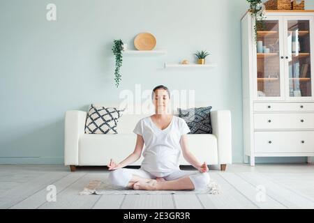 Lächelnde junge schwangere Dame in weiß meditiert im Padmasana sitzen Yogaposition auf dem Boden gegen Designermöbel im geräumigen Zimmer Zu Hause Stockfoto