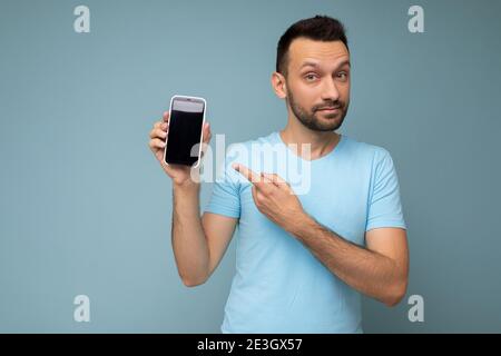 Schöner junger unrasiert Brunet Mann trägt jeden Tag blau T-Shirt isoliert Auf blauem Hintergrund halten und zeigen Handy mit leer Anzeige für Stockfoto