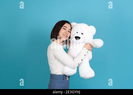 Überglücklich, gutherzig asiatische Frau steht seitlich und kuschelt sich zu einem großen weißen Teddybär isoliert auf einem blauen Hintergrund Stockfoto