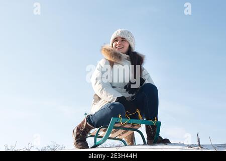 Frau sitzt und Schlitten vom Berg vor dem Hintergrund von Schnee und Himmel im Winter. Mädchen lacht und freut sich im Schnee, Unterhaltung im Schlitten Stockfoto