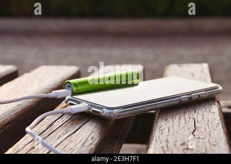 Smartphone und externe Powerbank liegen während des Ladevorgangs auf einer Holzbank im Park. Stockfoto