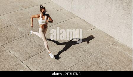 High-Angle-Ansicht einer Frau trägt Gesichtsmaske sprintet im Freien. Weibliche Laufen Training im Freien trägt eine schützende Gesichtsmaske. Stockfoto