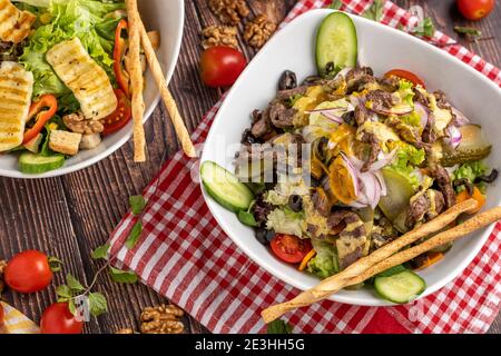 Halloumi-Salat und Rindfleischsalat in zwei separaten Tellern auf Holzhintergrund. Stockfoto