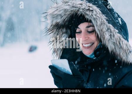Hübsche junge Frau im Wintermantel, Hut und Handschuhe lächelt, während sie ihr Handy mitten im Schneefall anschaut Stockfoto