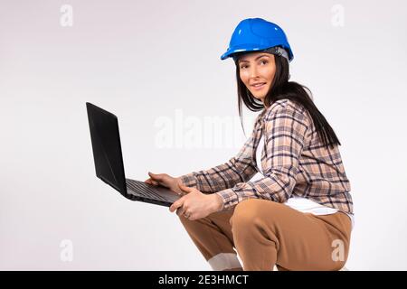 Eine Frau in einem blauen Helm posiert mit einem Laptop auf weißem Hintergrund und blickt mit einem Lächeln in die Kamera. Stockfoto