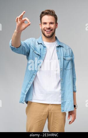 Porträt von jungen schönen kaukasischen Mann in Jeans Hemd zeigen ok Zeichen Geste stehen über hellen Hintergrund Stockfoto