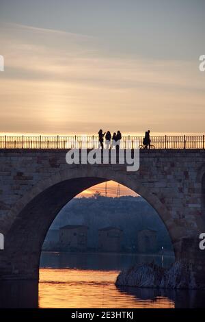 Menschen, die bei Sonnenuntergang auf einer Steinbrücke spazieren und die gefrorenen Bäume im Winter zur goldenen Stunde. Steinbögen, Spiegelung der Sonne auf dem Fluss. Abwasserkanäle Stockfoto