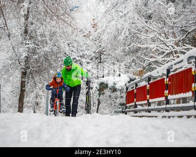 Vater lehrt seinen kleinen Sohn, im Winter Fahrrad zu fahren. Ein aktiver Zeitvertreib an der frischen Luft nach einem Schneefall in einem Winterpark. Stockfoto