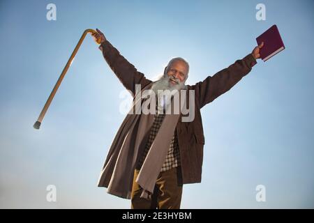 EIN BÄRTIGER ALTER MANN, DER DIE KAMERA ANSCHAUT, WÄHREND ER GLÜCKLICH AUFSTEHT HÄNDE Stockfoto