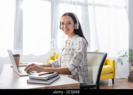 Fröhlicher Freiberufler mit Kopfhörern, der beim Tippen die Kamera anlächelt Notebook Stockfoto