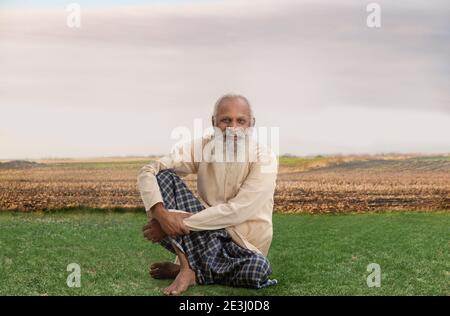 EIN GLÜCKLICHER ALTER MANN SITZT AUF GRAS MIT ACKERLAND IN HINTERGRUND Stockfoto