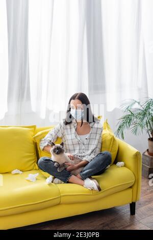 Junge allergische Frau in medizinische Maske leiden an Allergie, während Sitzend auf dem Sofa mit Katze und zerknitterten Servietten Stockfoto