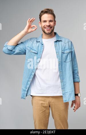 Porträt von jungen schönen kaukasischen Mann in Jeans Hemd zeigen ok Zeichen Geste stehen über hellen Hintergrund Stockfoto