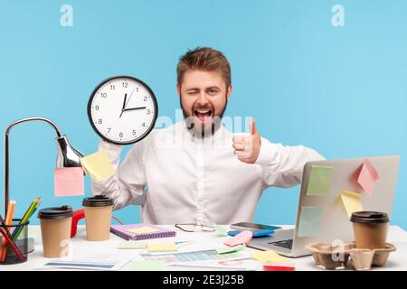 Glücklich lächelnder Mann Büroangestellter mit Bart in weißem Hemd zeigt Daumen nach oben und zwinkend hält Wanduhr, Beendigung der Arbeit auf Zeit, Zeitmanagement. ICH Stockfoto