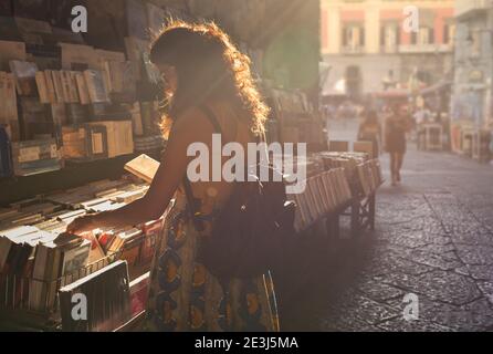 Eine junge Frau, die in Sommerkleidung gekleidet ist, wählt ein Buch, während sie auf einem europäischen Markt unter freiem Himmel einkaufen. Stockfoto